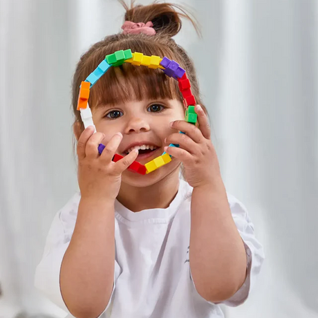 A smiling child with bangs and a ponytail holds a colorful ring of connected plastic blocks, looking playful and joyful against a soft background.