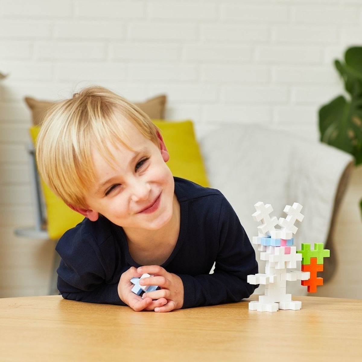 Child smiling next to a white bunny figure and carrot made from Plus-Plus building blocks on a wooden table.
