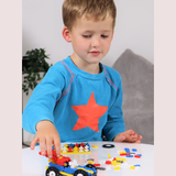 A young child in a blue sweatshirt with an orange star plays with building blocks on a table. Focused expression, blocks include colorful pieces and a small car.