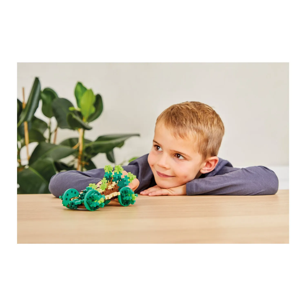 A young boy smiles while playing with a colorful toy car on a table, surrounded by greenery. The scene conveys creativity and joy.