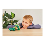 A young boy smiles while playing with a colorful toy car on a table, surrounded by greenery. The scene conveys creativity and joy.