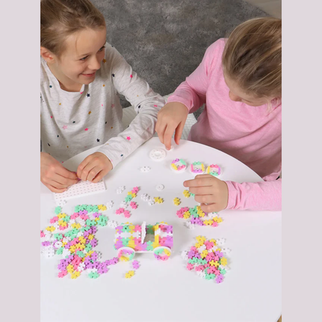 Two children happily play at a white table with colorful plastic building pieces, creating a small vehicle. The scene conveys creativity and joy.