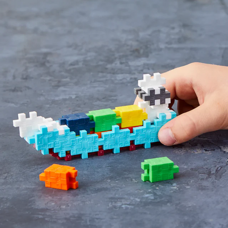 A hand holding an abstract boat model made of colorful interlocking puzzle pieces on a gray surface. Loose pieces, orange and green, lie nearby.