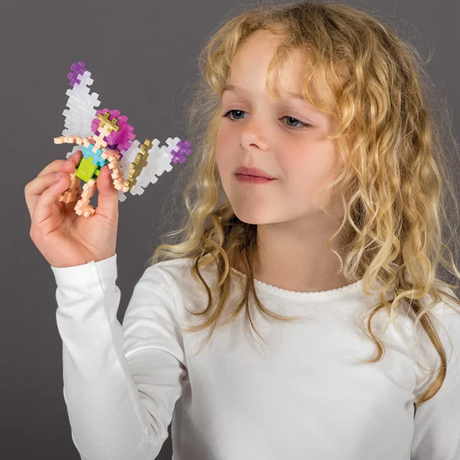 A young girl with curly blonde hair, wearing a white top, holds a colorful toy figure resembling a fairy with wings, showcasing curiosity and delight.