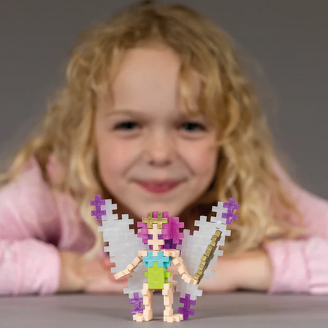 A young child with curly blond hair smiles, focusing on a colorful pixel-style fairy toy with pink hair and wings in the foreground, conveying joy.