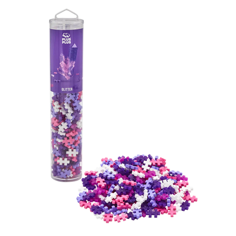 A clear tube labeled "Plus Plus Glitter" filled with pastel pink, purple, and white connecting pieces stands upright. A pile of pieces sits beside it.