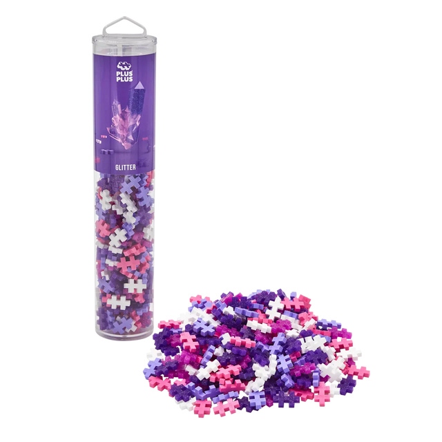 A clear tube labeled "Plus Plus Glitter" filled with pastel pink, purple, and white connecting pieces stands upright. A pile of pieces sits beside it.