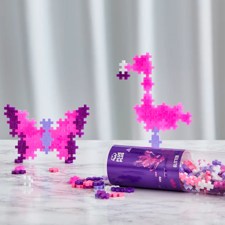 Colorful Plus-Plus blocks form a vibrant butterfly and flamingo. An open cylinder container lies on a marble surface, with blocks scattered around. Playful and creative mood.