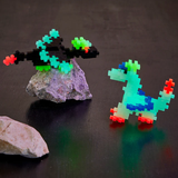 Glow-in-the-dark pixelated dinosaur and spaceship made of small, colorful interlocking pieces. The spaceship rests on a rock, set on a dark surface.