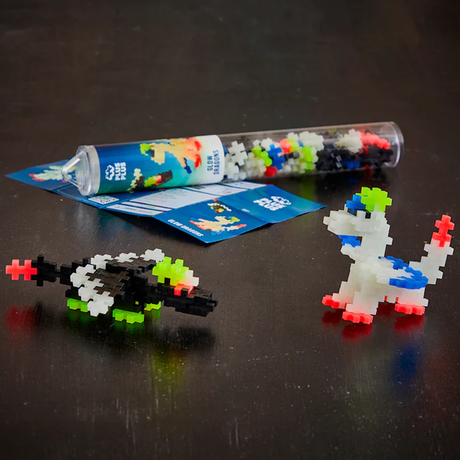 A black surface displays colorful Plus-Plus building pieces with two completed glow-in-the-dark dinosaur and tank models nearby. A plastic tube and instruction sheet lie behind them.