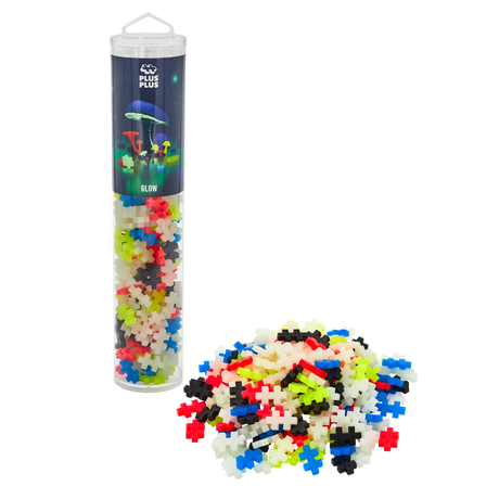 Tube of Plus-Plus Glow puzzle pieces with colorful interlocking pieces piled next to it. Packaging displays glowing mushrooms, creating a playful tone.