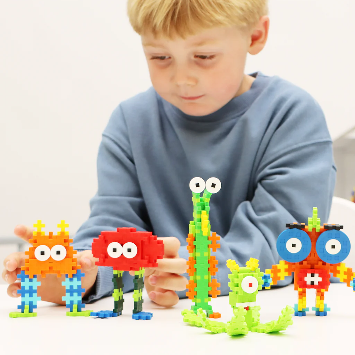 A young boy playing with colorful Plus-Plus toy creatures built with interlocking plastic blocks on a white table.