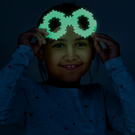 A smiling child wearing pixelated, glowing green glasses holds them up with both hands in a dark setting, creating a playful and fun atmosphere.