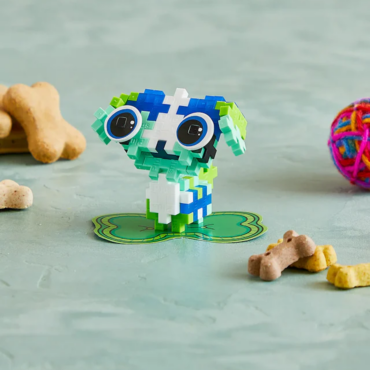 Colorful block toy resembling an animal stands on green base, surrounded by dog treats and a colorful ball on a light textured surface. Playful tone.