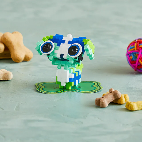Colorful block toy resembling an animal stands on green base, surrounded by dog treats and a colorful ball on a light textured surface. Playful tone.