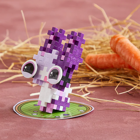 A purple and white pixelated bunny toy with large eyes stands on a green base. Straw and two carrots lie in the background on a pink surface.