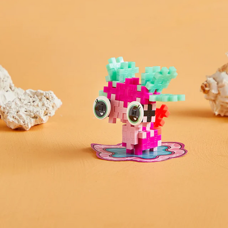 Colorful toy creature made of interlocking blocks on an orange surface, featuring large eyes and small wings. Coral pieces rest nearby, creating a playful scene.