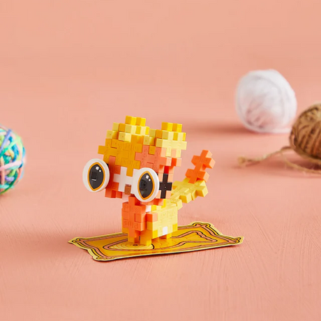 A colorful toy cat made of small interlocking blocks stands on an orange surface. It has large round eyes and is surrounded by balls of yarn.
