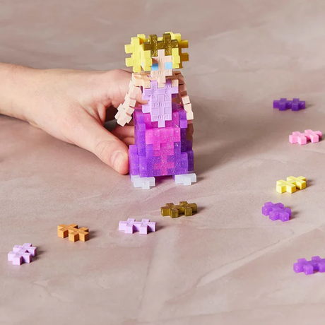 A hand holds a colorful pixelated princess figure made of interlocking blocks, in shades of pink, purple, and yellow, surrounded by scattered blocks.