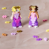 Two blocky princess figures made of interlocking pastel blocks, one with blonde hair and the other brown, stand on a textured surface with scattered blocks.