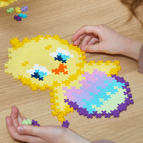 A child's hands assembling a yellow chick puzzle made of Plus-Plus blocks on a wooden table.