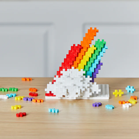 Colorful interlocking blocks form a rainbow emerging from white clouds on a wooden table. Scattered pieces add vibrancy and playfulness to the scene.