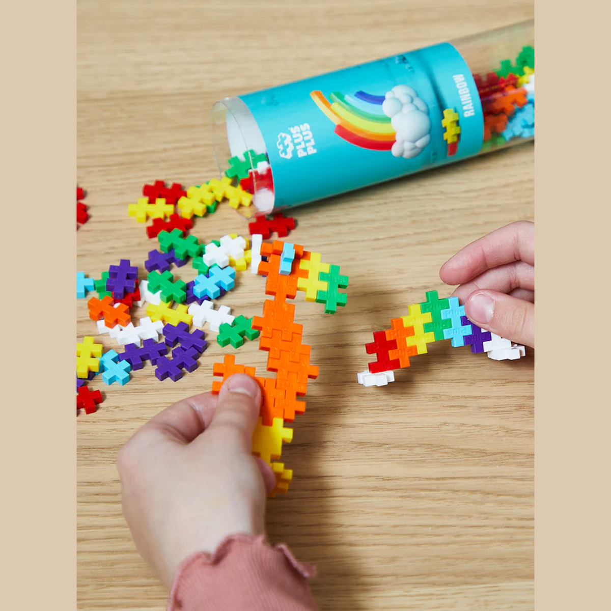 Hands assembling colorful interlocking plus-shaped pieces on a wooden surface. A transparent tube labeled “Plus Plus Rainbow” in the background. Playful and creative mood.