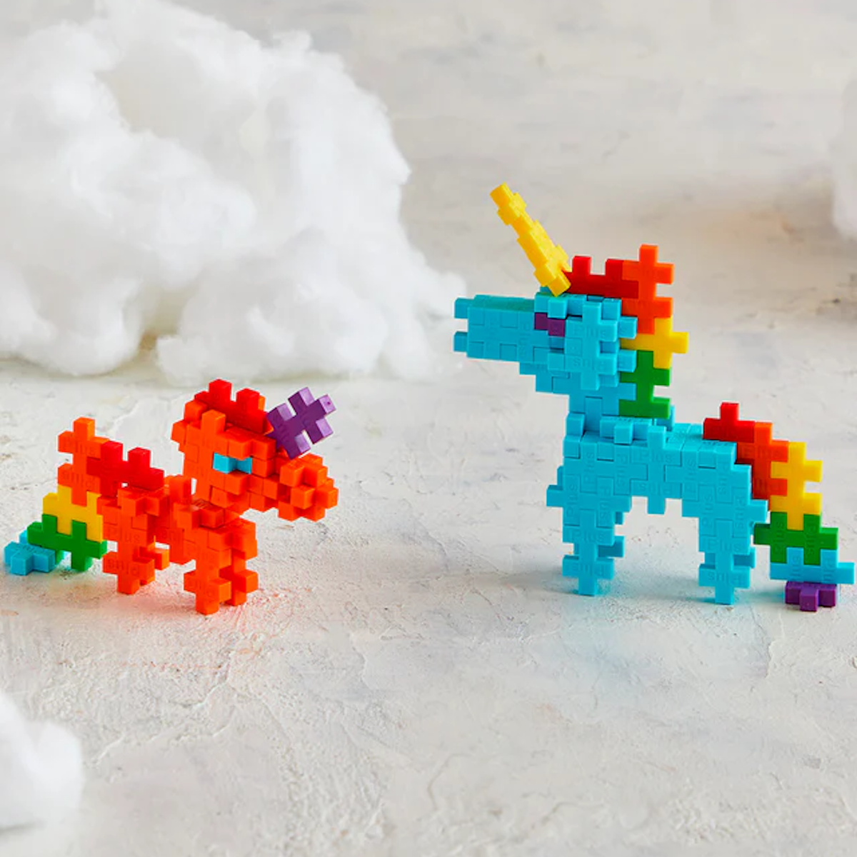 Toy unicorns built from colorful interlocking blocks: one blue with a yellow horn, the other orange with a purple horn, set against a cloud-like backdrop.