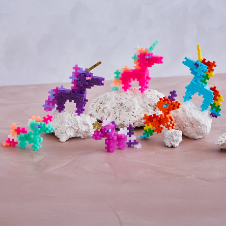 Colorful pixelated unicorn figures in vibrant pink, purple, blue, and orange, arranged on white stones against a pale pink surface. Playful and whimsical.