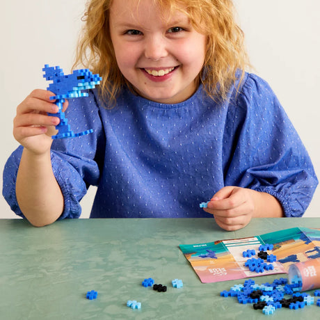 Smiling girl holding a blue Plus-Plus dolphin. Creative STEM building toy for kids with instruction manual.