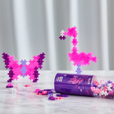 Pink glitter Plus-Plus blocks built into a butterfly and flamingo, with a storage tube on a marble surface.