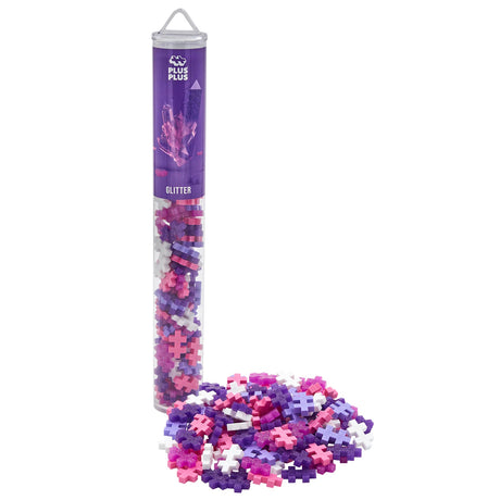 Plus-Plus Glitter building blocks in a clear tube with a pile of pink, purple, and white shimmering cross-shaped pieces.