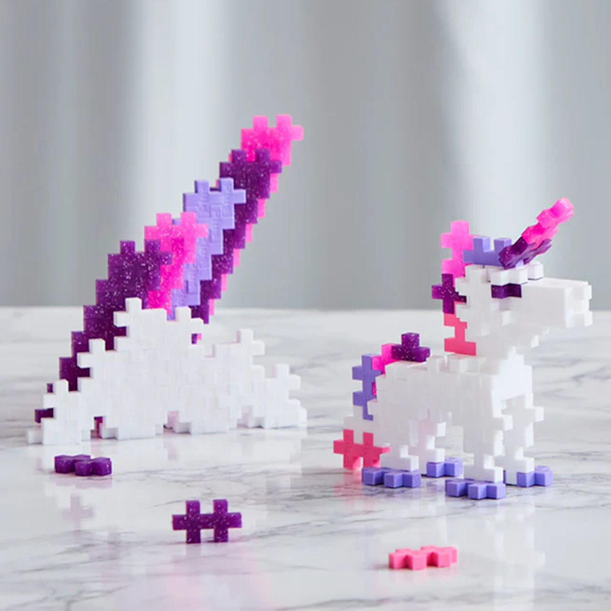 A white unicorn and mountain built from pink, purple, and white glitter Plus-Plus blocks on a marble surface.