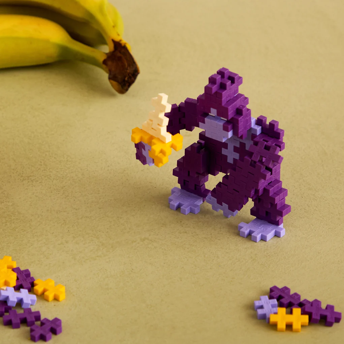 Purple gorilla figure built with Plus-Plus blocks, holding a banana near real bananas on a tan surface.