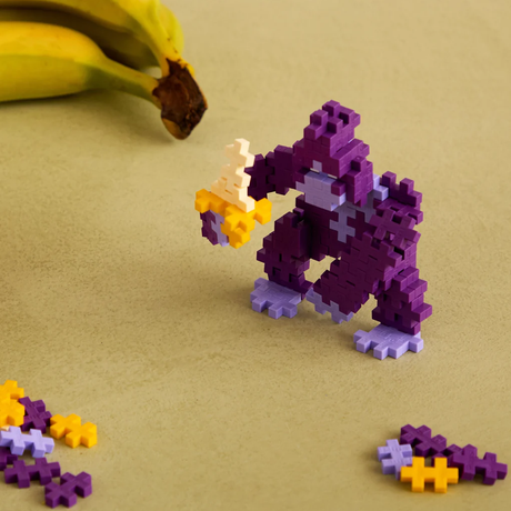 Purple gorilla figure built with Plus-Plus blocks, holding a banana near real bananas on a tan surface.