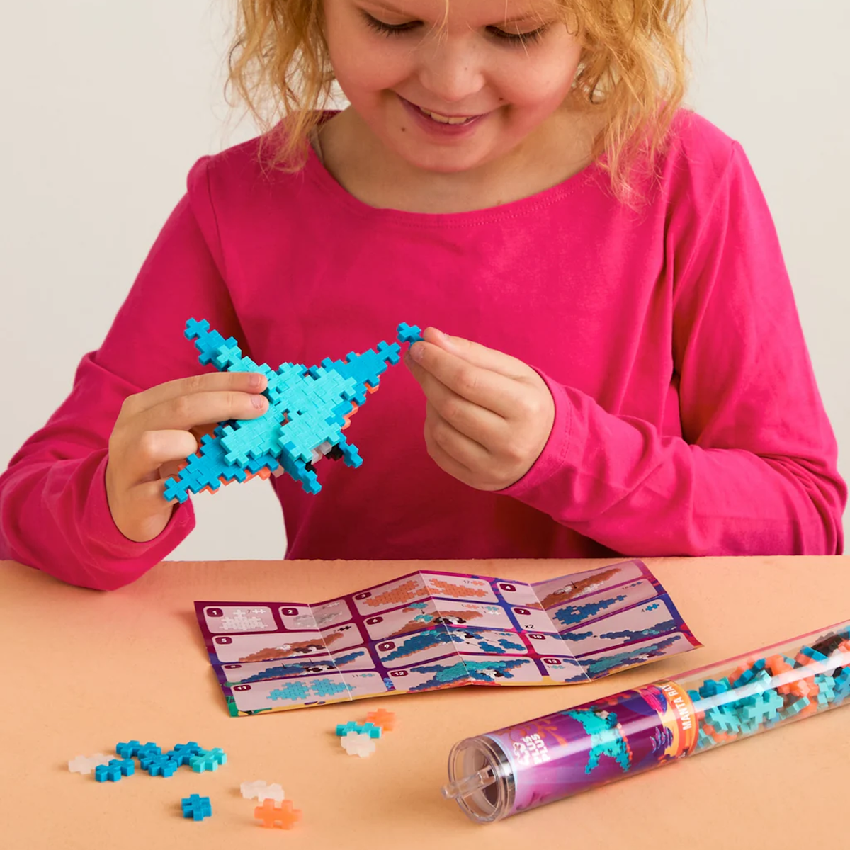 Child building a blue manta ray using Plus-Plus blocks, with instructions and a storage tube on a peach surface.