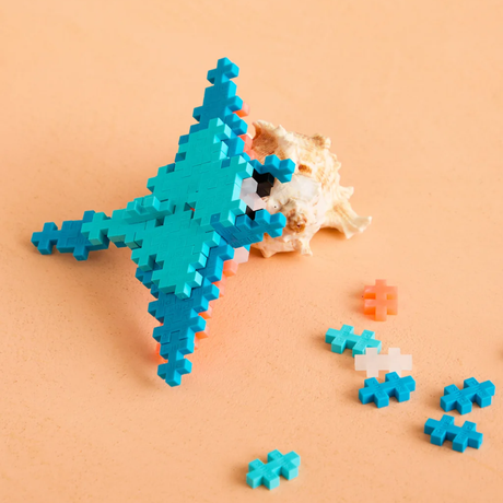 Plus-Plus blocks manta ray toy model next to a seashell on a sandy background, with scattered teal and orange pieces.