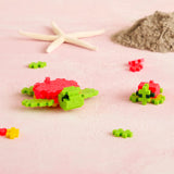 Plus-Plus sea turtles made with small green and red interlocking blocks. Building toy animals on a pink beach-themed surface.