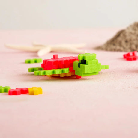 A lime green and coral pink sea turtle made of Plus-Plus blocks on a pink sandy surface with a starfish background.