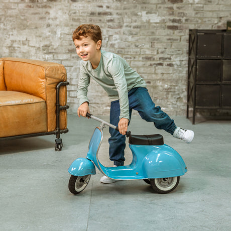 A smiling child with curly hair balances on a small blue scooter indoors. A brown leather couch and brick wall are in the background, creating a playful atmosphere.