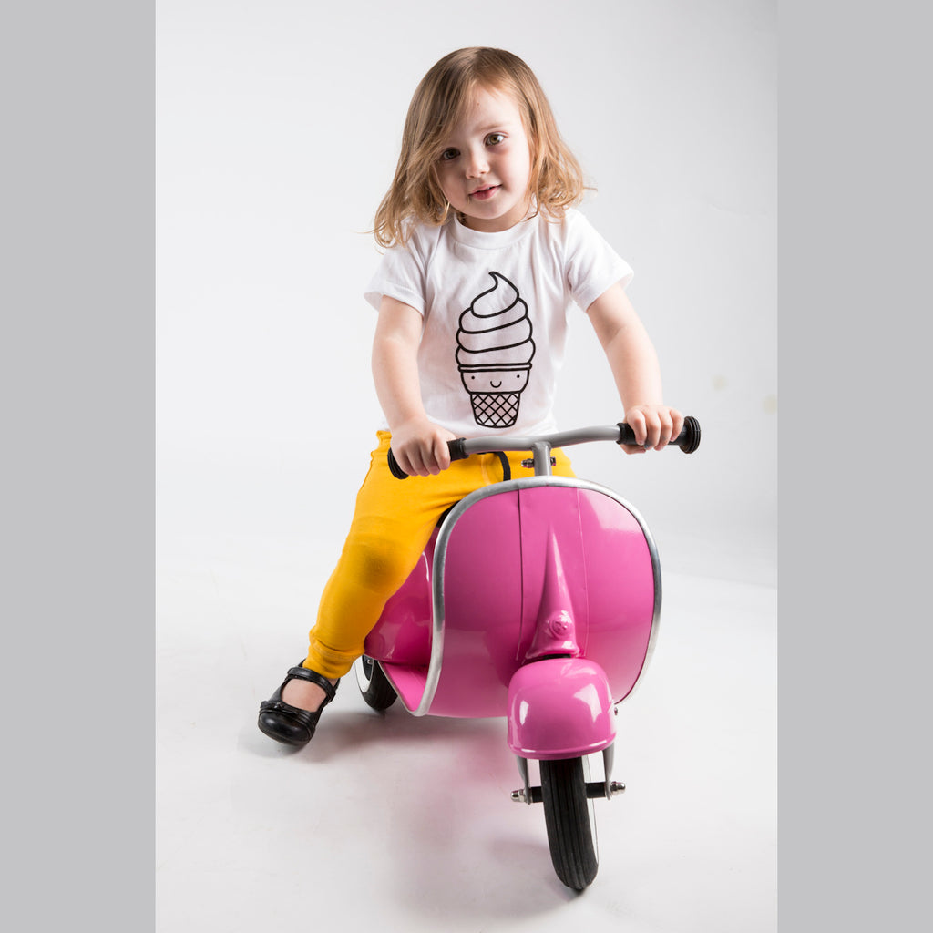 A young child with wavy hair smiles while sitting on a pink toy scooter. They wear a white ice cream print shirt, yellow pants, and black shoes.