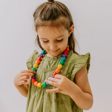 A young girl in a green dress smiles down at a colorful, chunky bead necklace she holds. The background is plain, emphasizing her curiosity and joy.