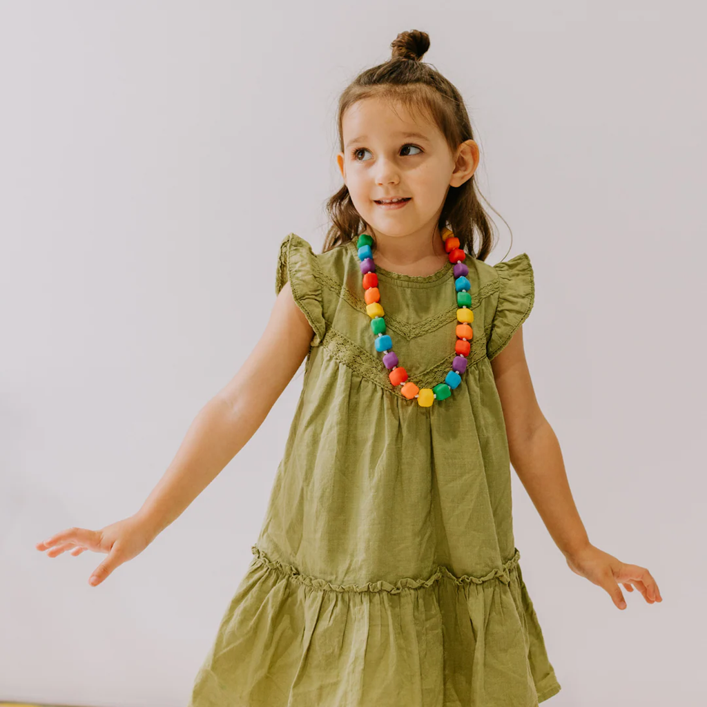 A young girl in a green dress and colorful bead necklace smiles softly against a plain background. Her hair is half-up, conveying a playful tone.