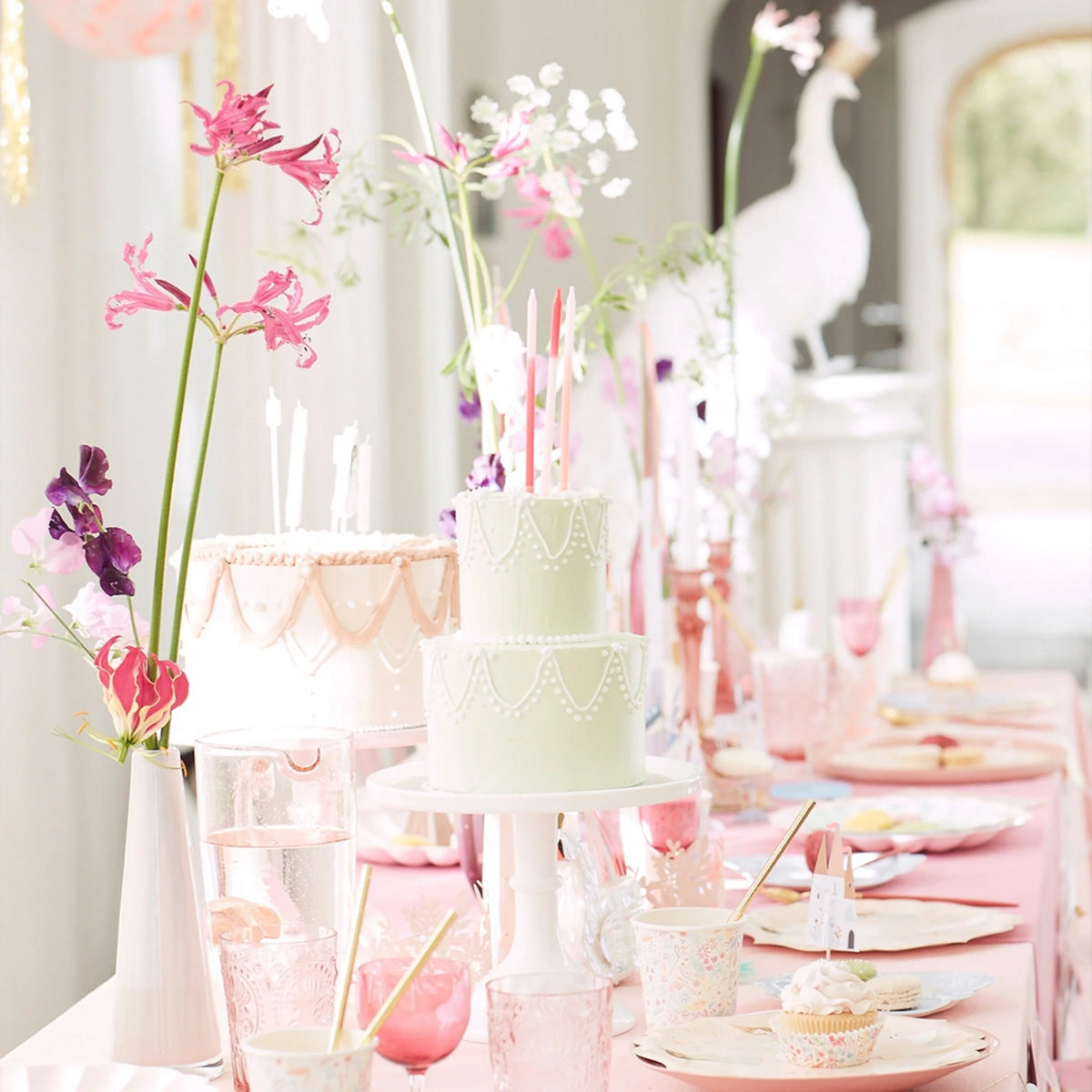 A beautifully decorated birthday party table with pastel cakes, pink flowers, and elegant tableware.