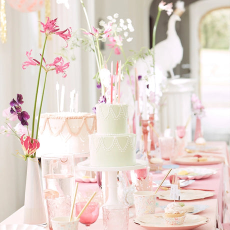 A beautifully decorated birthday party table with pastel cakes, pink flowers, and elegant tableware.
