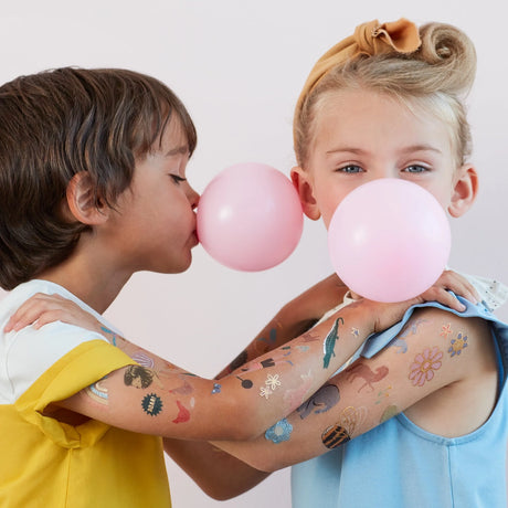 Two children with arms covered in colorful temporary tattoos each blow a large bubble with pink bubble gum.