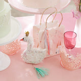 Pink party table with a swan piñata favors bag, small crowns, cakes, and a pink glass.