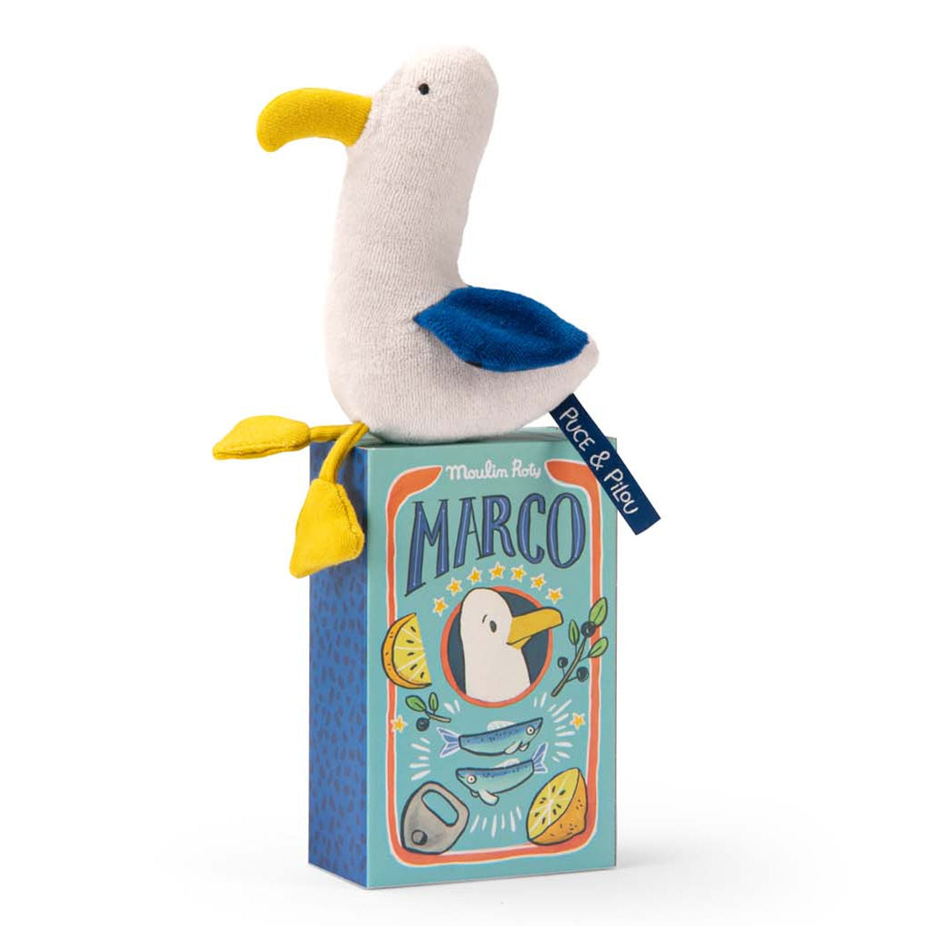 A plush seagull toy with a yellow beak and feet, blue wings, and a white body sits atop a colorful box labeled "Marco" with playful illustrations.