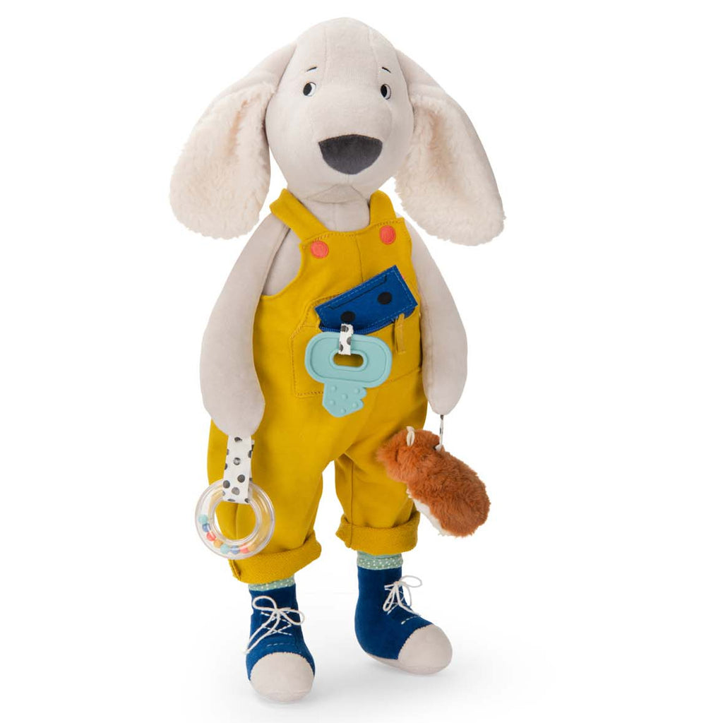 Plush dog toy wearing yellow overalls and blue shoes, holding a clear rattle and fuzzy brown accessory. The image conveys a playful and cheerful tone.