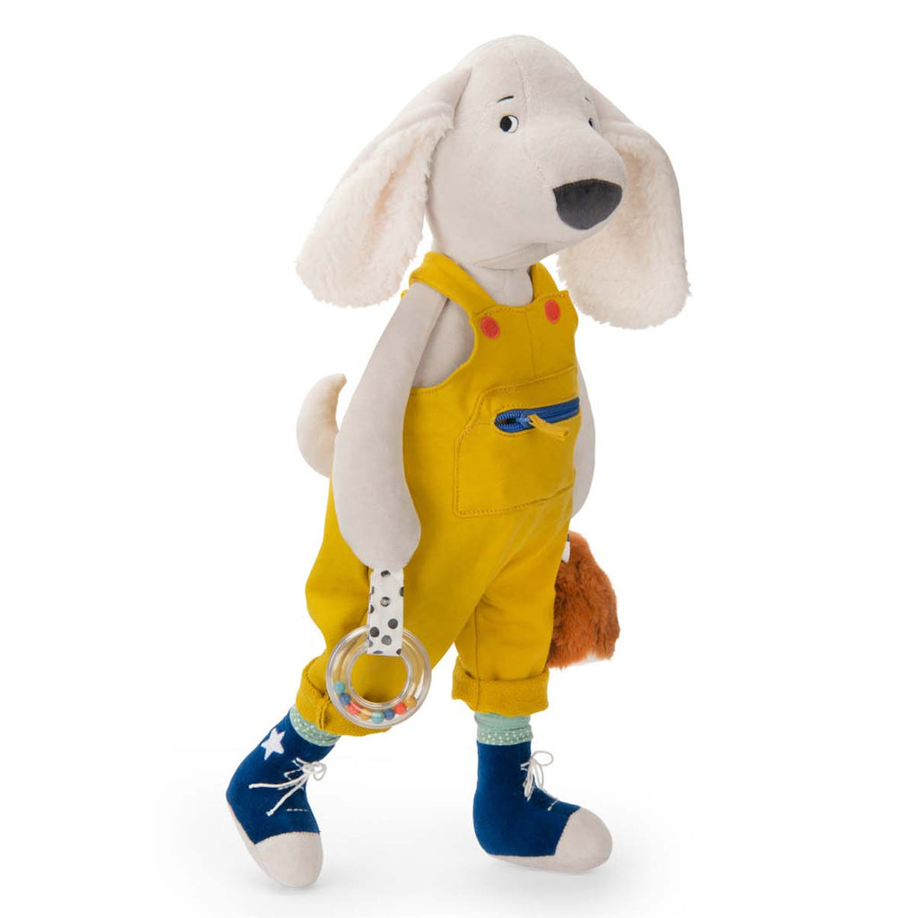 Plush dog toy wearing yellow overalls and blue shoes, holding a clear rattle and fuzzy brown accessory. The image conveys a playful and cheerful tone.